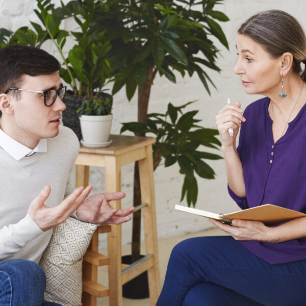 Psychology, therapy, psychiatry, mental health and counseling concept. Candid shot of nervous self conscious young male in glasses telling middle aged female counselor about his problems at work
